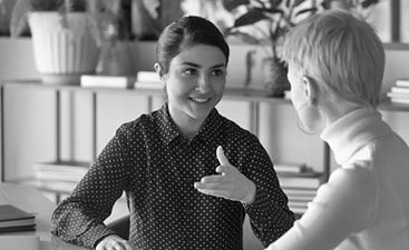 Friendly millennial indian businesswoman talking to blonde female colleague, sitting at table in office  Two young multiracial employees discussing working issues or enjoying informal conversation 